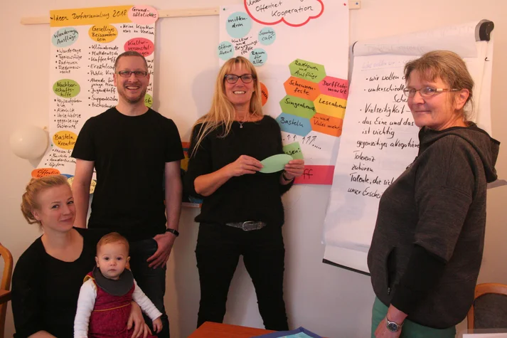 Drei Erwachsene stehen an einer Flipchart, im Vordergrund sitzt eine junge Frau mit einem Baby auf dem Schoß.