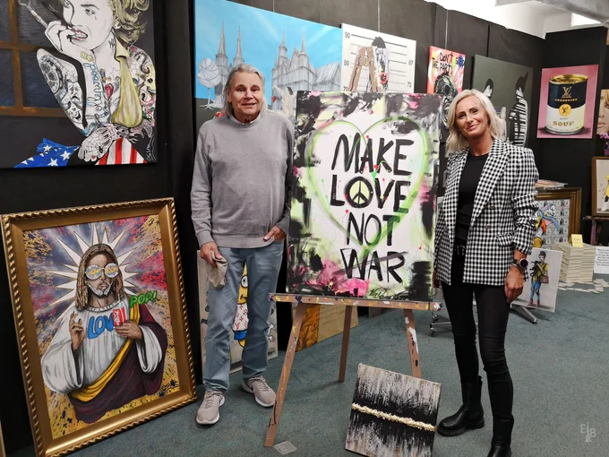 Oskar Müller und Ina Daume stehen links und rechts neben einem Kunstwerk mit der Aufschrift "Make Love not War", im Hintergrund sind weitere Werke zu sehen.