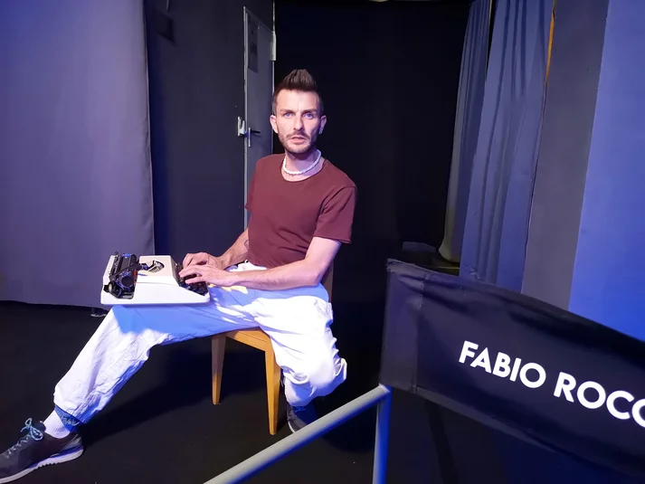 Auf einem Holzhocker sitzt ein Mann mit weißer Hose und rotem T-Shirt. Auf seinem Schoß ist eine weiße Schreibmaschine. Im Vordergrund ist ein schwarzer Regiestuhl mit weißer Aufschrift "Fabio Rocco"