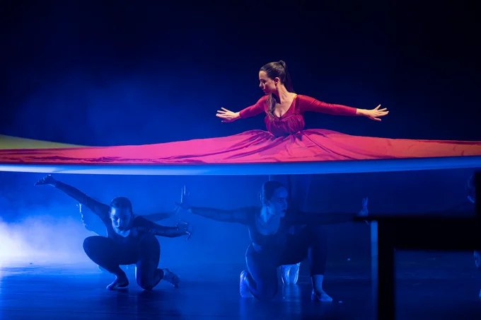Im oberen Bildteil ist der Oberkörper einer braunhaarigen Tänzerin im roten Kleid zu sehen. Das Kleid teilt das Bild und im unteren Bildteil sieht man die Beine der Tänzerin, die auf den Zehenspitzen steht sowie drei Tänzerinnen, die in schwarzen Kostümen genügt auf der Bühne sind
