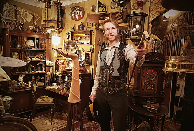 Dr. Wolf begrüßt seine Gäste in der Wunderkammer – er streckt eine Hand mit Gehstock in Richtung Kamera. Links neben ihm ist eine Nachbildung eines Arms zu sehen, die eine Glaskugel balanciert.