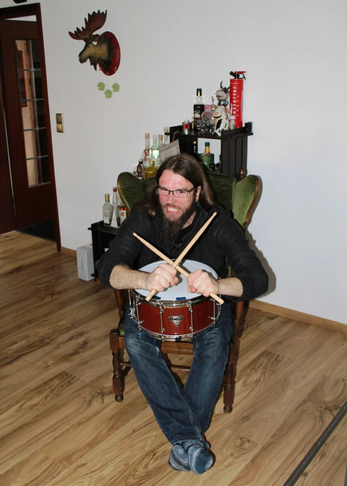 Niklas Kahl posiert mit gekreuzten Drumsticks und grimmiger Miene für die Kamera. Er sitzt in einem Ohrensessel in seinem Wohnzimmer.