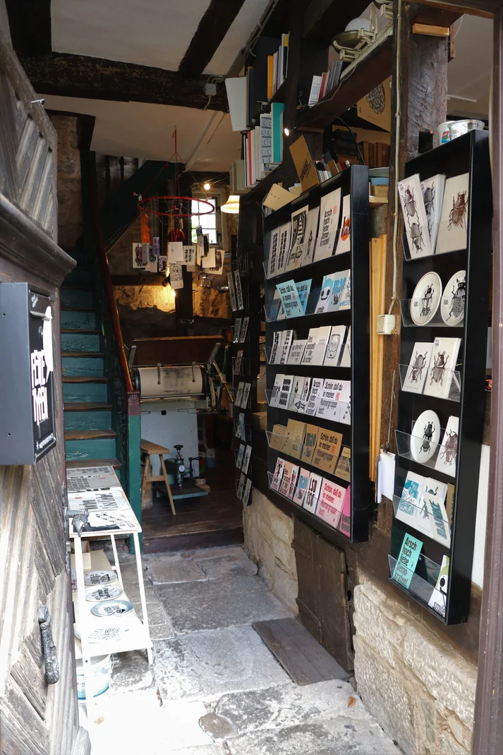 Eine alte Druckerei in einem Fachwerkhaus. An den Seiten sind bedruckte Postkarten. Im Hintergrund ist eine hellblaue Treppe zu sehen.