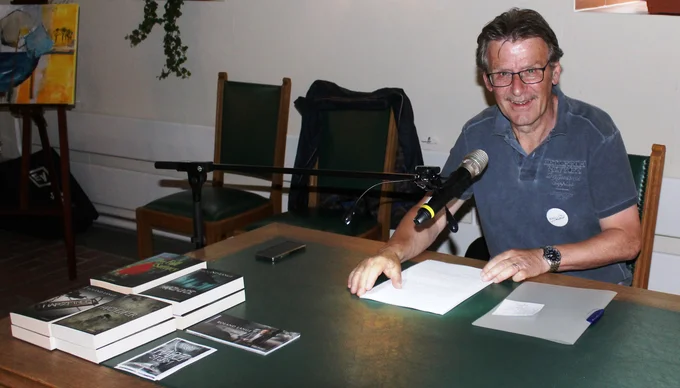 Roland Lange sitzt lächelnd an einem Schreibtisch, vor ihm ein aufgeschlagenes Buch und ein Mikrofon.