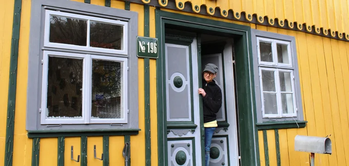 Die Künstlerin Kerstin Schulz schaut aus der Tür eines gelben Hauses hinaus mit grünen Streben, in denen Bleistifte eingearbeitet sind