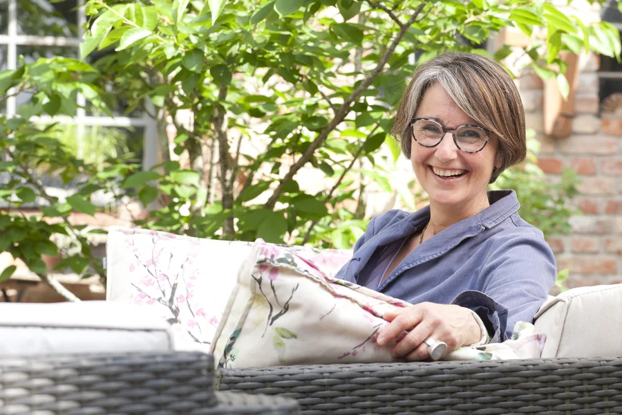 Anja Fehrensen lacht in die Kamera, sie sitzt auf einem Sessel im freien, im HIntergrund grüne Blätter.