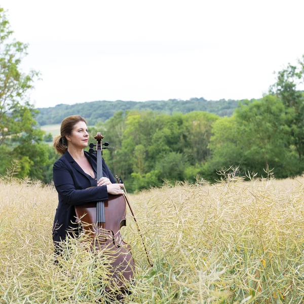 Lucile Chaubard steht mit einem Cello in einem Rapsfeld, im Hintergrund erstreckt sich ein waldiger Hügel.