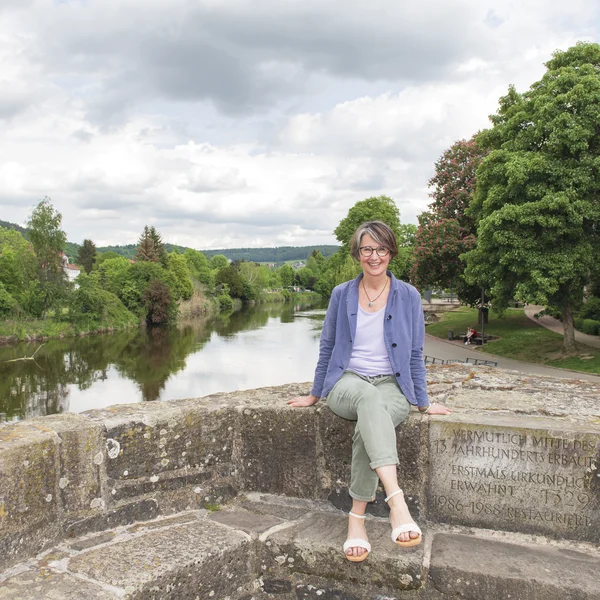 Anja Fehrensen sitzt lächelnd auf einer Steinmauer, im Hintergrund sieht man einen Flusslauf mit grünen Bäumen.