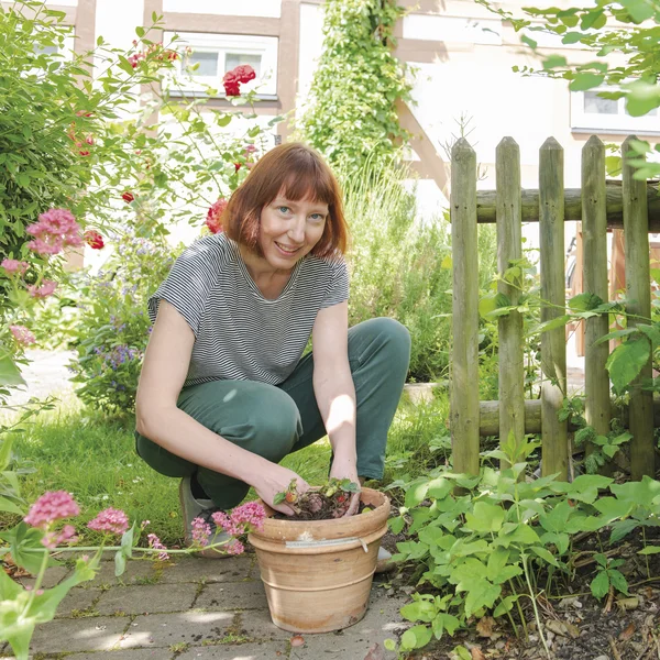 Ruth Reiche kniet in einem Garten und lächelt in die Kamera, ihre Hände sind voller Erde und stecken in einem Blumentopf mit Erdbeeren.