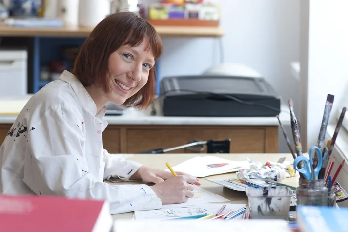 Ruth Reiche sitzt im Malerkittel zeichnend an einem Schreibtisch und lächelt in die Kamera