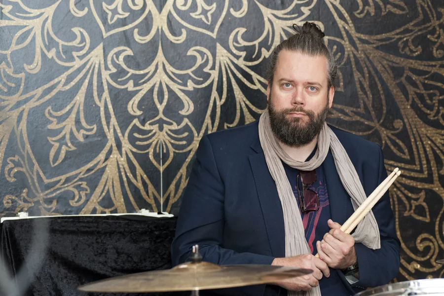 Tobias Langer sitzt vor einer gemusterten Tapete, in der Hand zwei Schlagzeugsticks, vor ihm ein Schlagzeug-Becken.