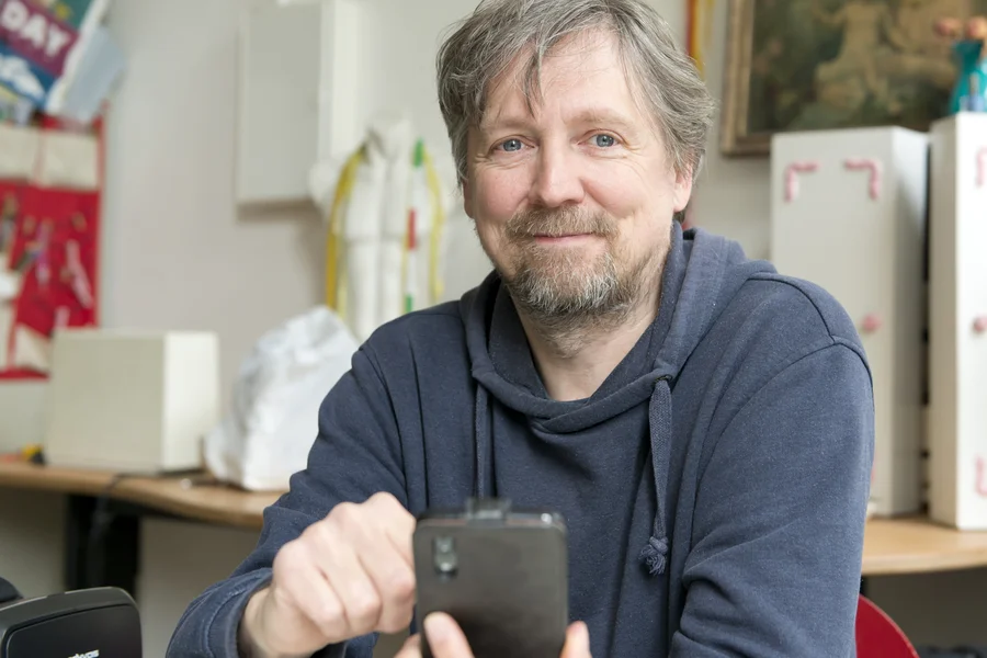 Thomas Kirchberg (alias Tokio) lächelt in die Kamera, in seiner Hand ein Smartphone