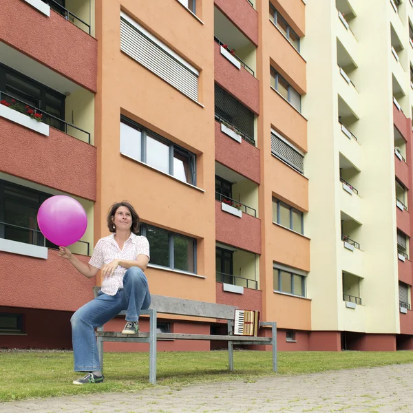 Norika Wacker sitzt mit einem rosa Ballon in der Hand auf der Lehne einer Bank, im HIntergrund die Balkone eines großen mehrstöckigen Wohnblocks.