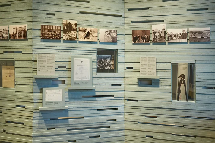 Wand aus Holzstäben mit eingebrachten Freiräumen für Exponate, zum Beispiel einem Bolzenschneider, der in den 1950er-Jahren genutzt wurde, um während einer Flucht den Stacheldraht durchzutrennen.