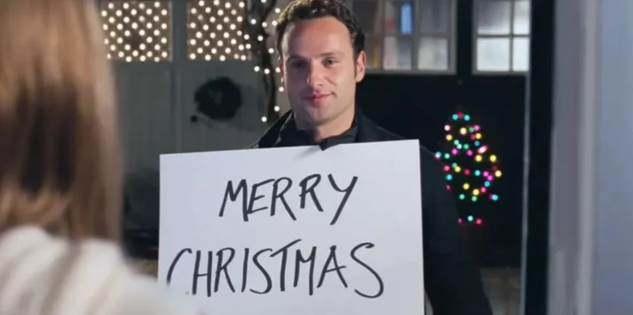 Andrew Lincoln zeigt Keira Knightely ein Schild auf dem frohe Weihnachten steht.