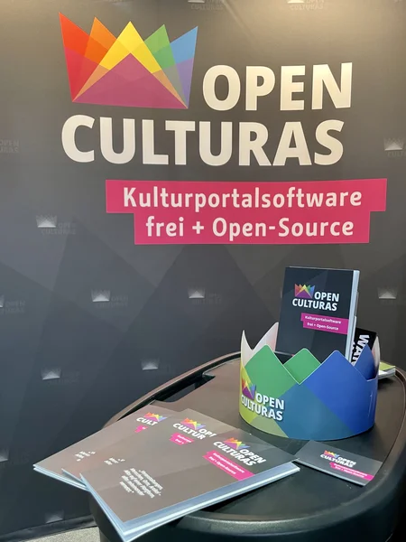 OpenCulturas-Messestand: im Hintergrund ein Banner mit der Aufschrift "OpenCulturas Kulturportalspftware frei + Open-Source", im Vordergrund Flyer, Visitenkarten und eine farbige Papp-Krone auf einem Tisch.