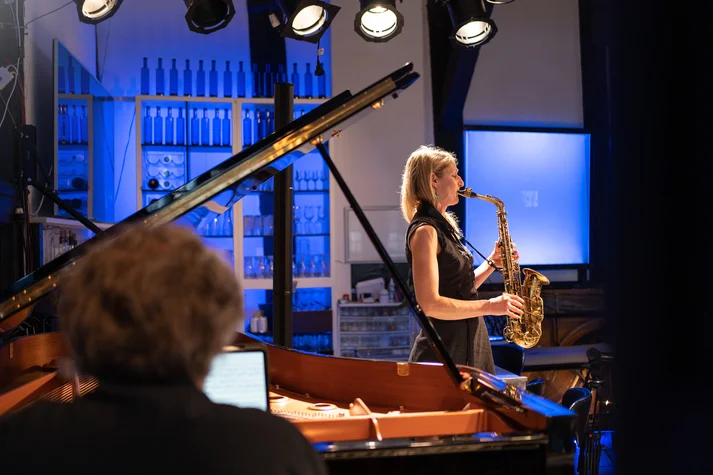 Eine Frau mit blonden haaren und schwarzem Kleid spielt Saxofon. Im Vordergrund sitz ein Mann mit grauen Haaren an einem Flügel.Michael Mehle