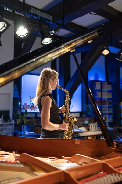 Eine Frau mit blonden haaren und schwarzem Kleid spielt Saxofon. Ein offener Flügel ist im Vordergrund