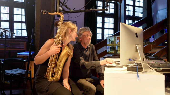 Eine blonde Frau im schwarzen Kleid mit Saxofon sitzt neben einem Mann im schwarzen Hemd. Beide schauen auf einen iMac