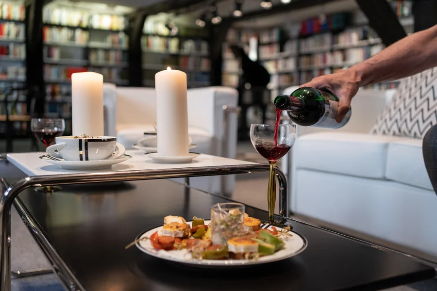 Ein schwarzer Couchtisch mit einer weißen Couch rechts daneben. Ein weißer Teller mit Essen und ein Weinglas, in das Rotwein eingegoßen wird stehen im Vordergrund. Auf dem Tisch sind zwei weiße Kerzen und zwei weiße Kaffeetassen sowie ein weiteres Glas Rotwein. Im Hintergrund ist ein weißer Couchsessel sowie Bücherregale zu sehen.