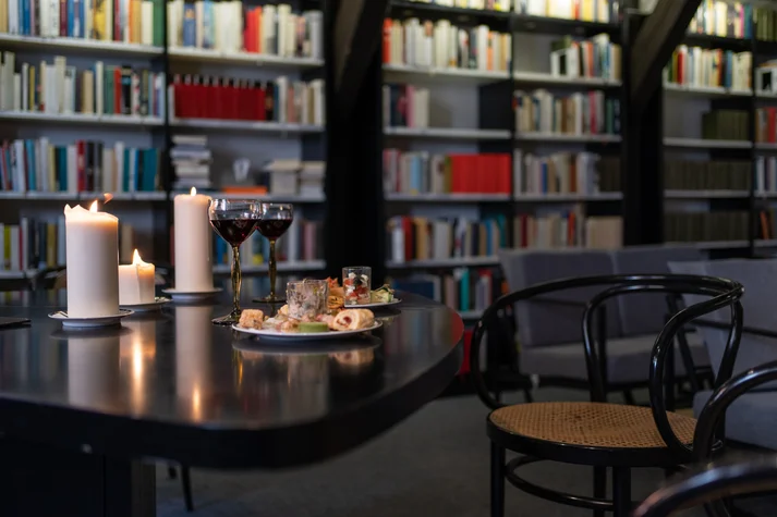 Ein schwarzer Tisch mit zwei Tellern Essen, zwei Rotweingläsern und drei brennenden weißen Kerzen. Rechts daneben sind zwei schwarze Stühle und im Hintergrund Bücherregale und eine weiße Couch