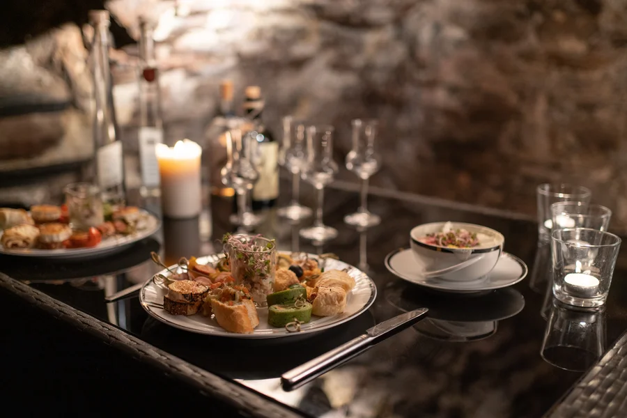 Zwei Teller Essen auf einem schwarzen Tisch mit verschiedenen Gläsern, Kerzen und einer Kaffeetasse in einem Kellergewölbe