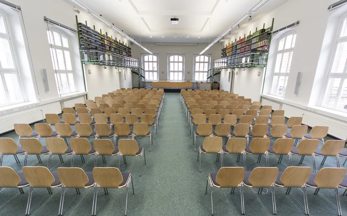 Der Alfred Hessel Saal im Historischen Gebäude der Händlerlogo Niedersächsische Staats- und Universitätsbibliothek Göttingen. Zu sehen sind Stuhlreihen, weiße Fenster an den Seiten und am Ende und hochhängende grüne Bücherregale.