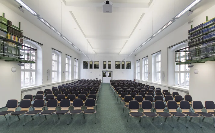 Der Alfred Hessel Saal im Historischen Gebäude der Händlerlogo Niedersächsische Staats- und Universitätsbibliothek Göttingen. Zu sehen sind Stuhlreihen, weiße Fenster an den Seiten und am Ende eine Tür sowie hochhängende grüne Bücherregale rechts und links.