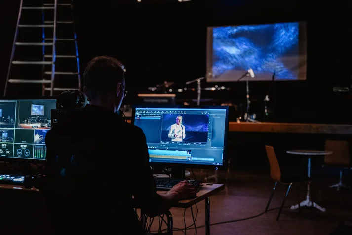 In einem dunklen Veranstaltungsraum sitzt ein Mann vor zwei bläulich leuchtenden Bildschirmen. Auf der dahinter liegenden Bühne ist eine Leinwand mit bläulichen Animationen zu sehen.