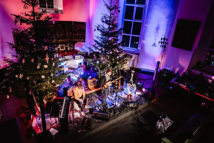 In einem rot-bläulich beleuchtetem Kirchenschiff am Altar stehen zwei Weihnachtsbäume. Davor aufgebaut ist eine dreiköpfige Band mit Gitarre, Cajòn und Schlagzeug. Ein Klavier steht ebenfalls auf der Bühne und rechts davon sind Streamingpulte aufgebaut, um die Veranstaltung aufzunehmen. 
