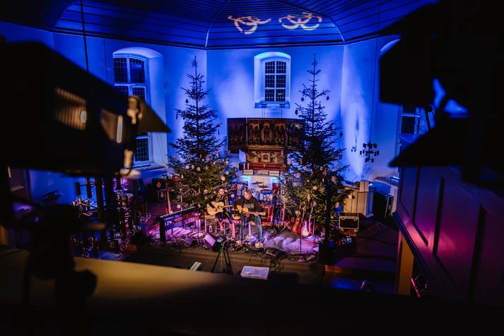 In einem rot-bläulich beleuchtetem Kirchenschiff am Altar stehen zwei Weihnachtsbäume. Davor aufgebaut ist eine zweiküpfige Band.