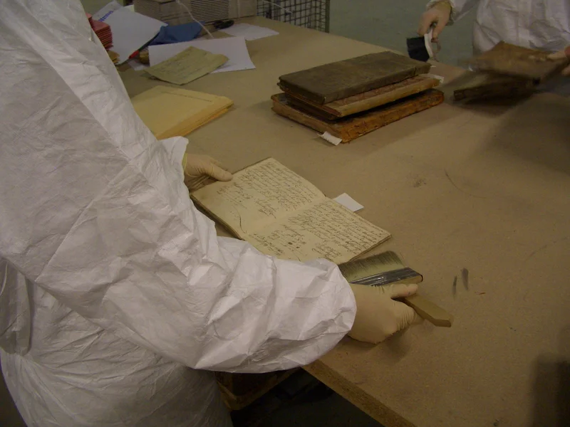 Eine Archivalie liegt auf einem Holzbrett, eine Person in weißer Schutzkleidung streicht die offenen Seiten mit einem Pinsel ab.