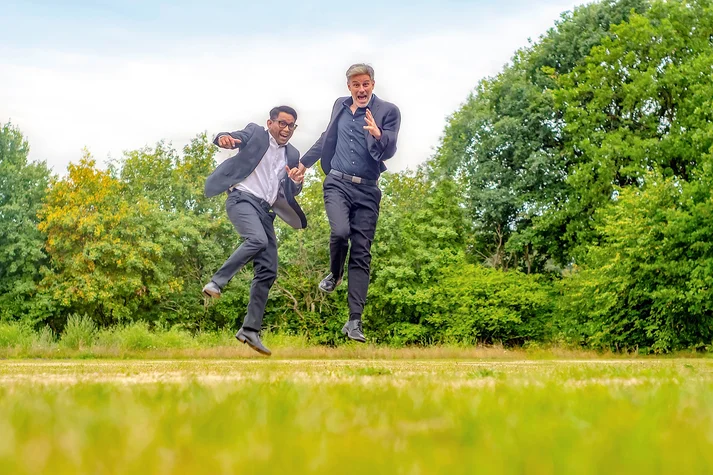 Zwei Männer im Anzug springen in die Luft, im Hintergrund grüne Landschaft.