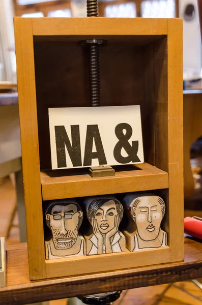 Eine weiße Postkarte mit der Aufschrift "NA &" steht in einer Holzpresse. in der Presse sind drei gezeichnete Köpfe.
