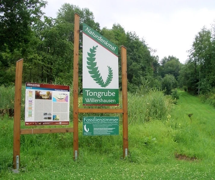 Infoschilder am Eingang zu Tongrube in grüner Landschaft; links zum Geopark Harz, rechts zur Tongrube Willershausen mit großer Farnmyrte als Logo