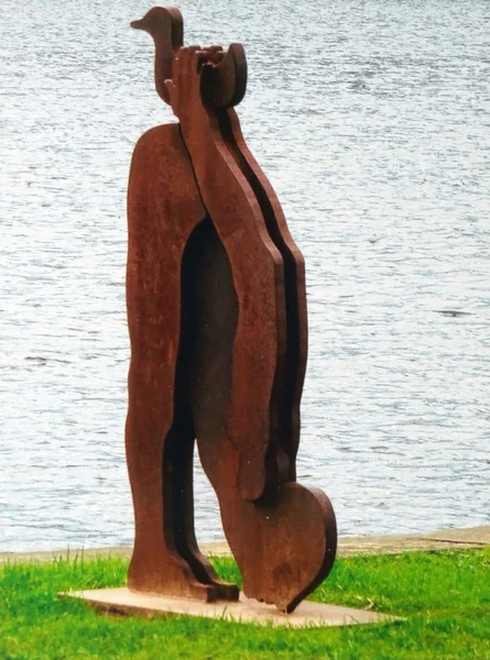 Stahl-Skulptur von Karl Repfennig: Eine Figur beugt sich auf den Boden, über ihrem Gesäß hält sie eine Ente. Im Hintergrund ein See.