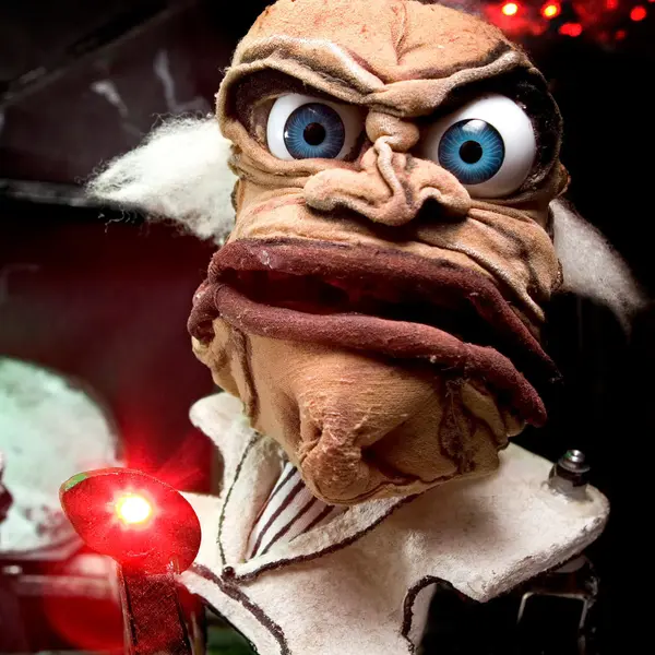 Nahaufnahme einer grotesk wirkenden Puppenfigur mit großen, starr blickenden blauen Augen, wulstigen Lippen und zerzausten weißen Haarsträhnen. Vor ihr leuchtet ein kleines rotes Licht.