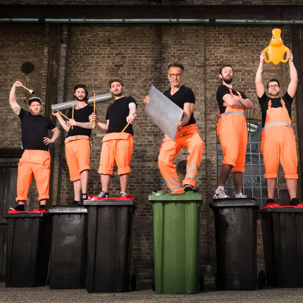 Sechs Männer in orangefarbenen Arbeitslatzhosen stehen auf schwarzen und einer grünen Mülltonne vor einer Backsteinwand. Sie halten verschiedene Alltagsgegenstände als Schlaginstrumente – Rohre, Drumsticks, ein Metallblech. Einer hält eine orangefarbene Gummifigur in die Höhe.