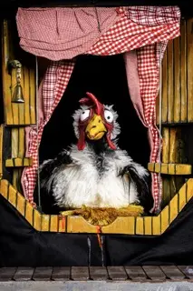 Huhn-Puppe mit aufgerissenen Augen schaut aus einem rustikalen Holzfenster mit rot-weißem Karostoff-Vorhang.