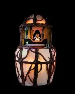 Beleuchtetes Theaterminiaturobjekt: eine eiförmige Konstruktion aus Ästen, im Inneren eine kleine Bühne mit Figur hinter rotem Vorhang. Schwarzer Hintergrund.