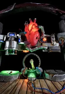 Theatermaschinerie: Ein leuchtendes rotes Herz hängt über einer grünen Schlangenfigur in einem Metallring, umgeben von technischen Apparaturen auf einer Holzbühne.