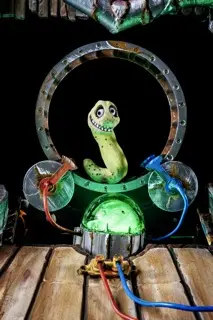 Grüne Schlangenfigur mit grinsendem Gesicht in einem beleuchteten Metallring auf einer Holzbühne, verbunden mit bunten Kabeln.
