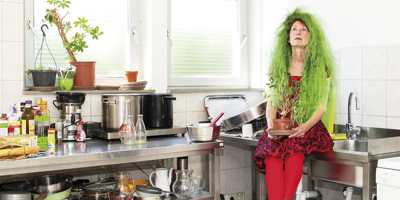 Franziska Aeschlimann sitzt auf einer Edelstahlspüle in einer Küche neben einem vollgestellten Etagenwagen. In der Hand hält sie eine Zimmerpflanze auf dem Kopf hat sie eine Perücke mit langen wilden grünen Haaren.