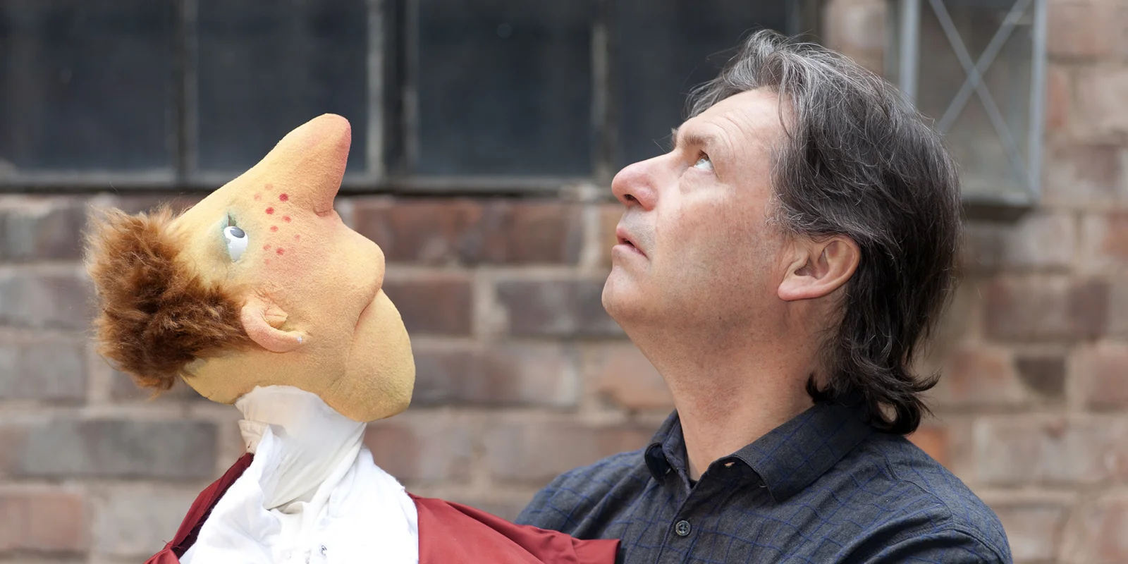 Christoph Buchfink blickt in den Himmel, auf seiner Hand hat er eine Puppe, die es ihm gleich tut. Im Hintergrund ist eine Steinmauer mit alten Fenstern und einer Laterne.