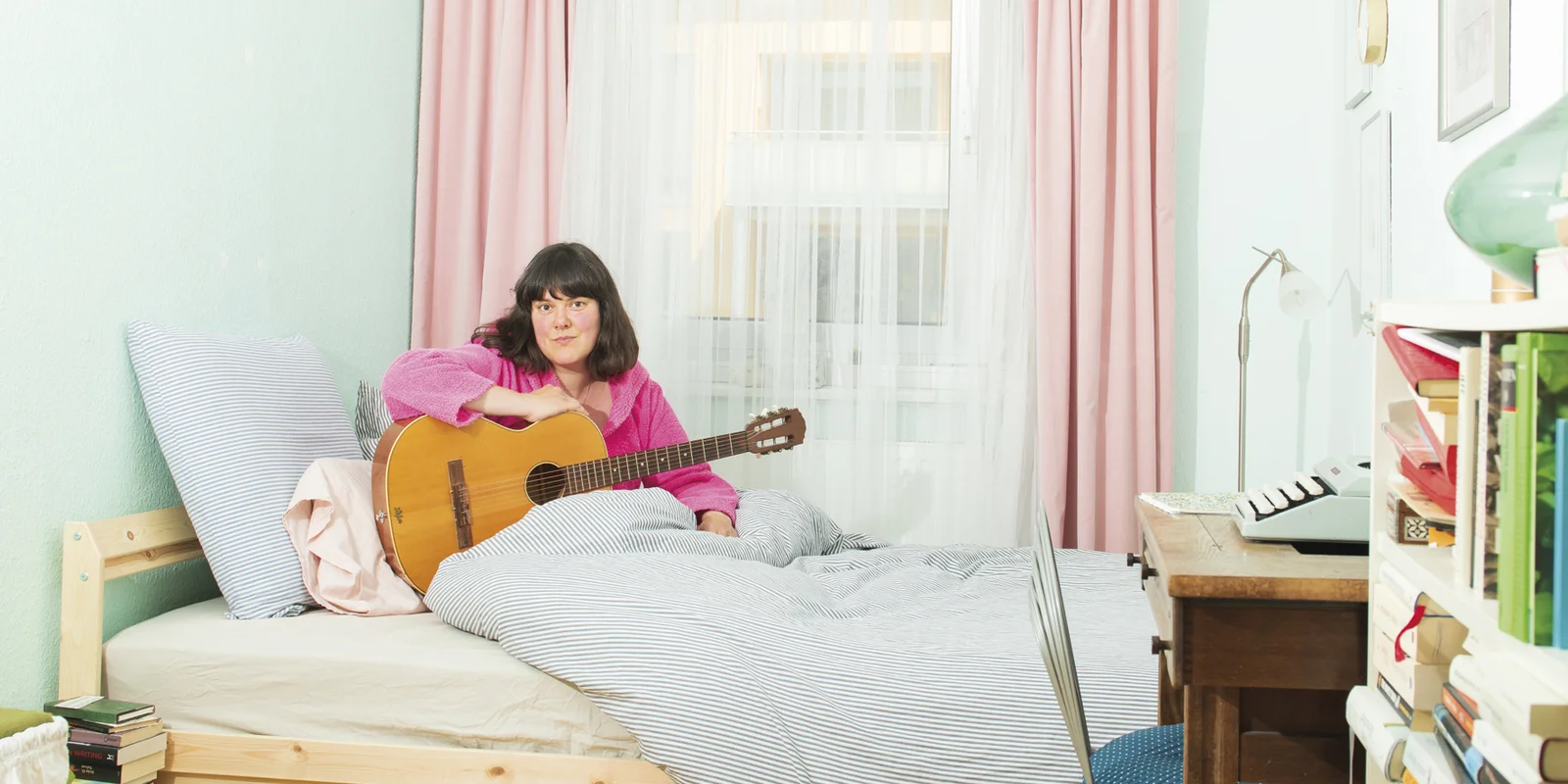 Frau Pauli sitzt im rosa Bademantel auf einem Bett, eine Gitarre vor sich, auf die sie ihren rechten Arm abgelegt hat.