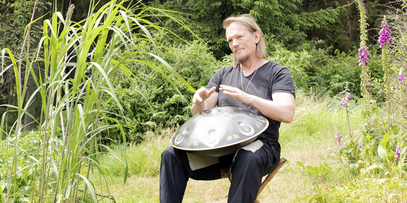 Rolf Predotka (alsia Hangklang) spielt auf einem Stuhl sitzend auf einer Hangdrum. Im Hintergrund ist ein grüner Garten zu sehen.