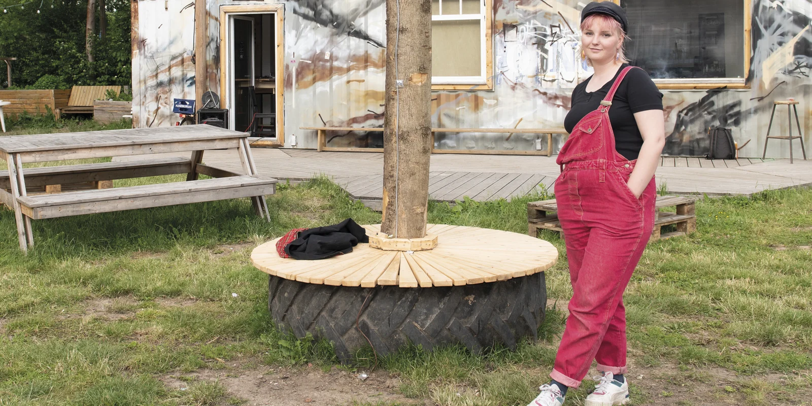 Marlin Helene steht mit überkreuzten Beinen vor einem mit Grafitti besprühtem Container, danaeben mehrere Bäume.