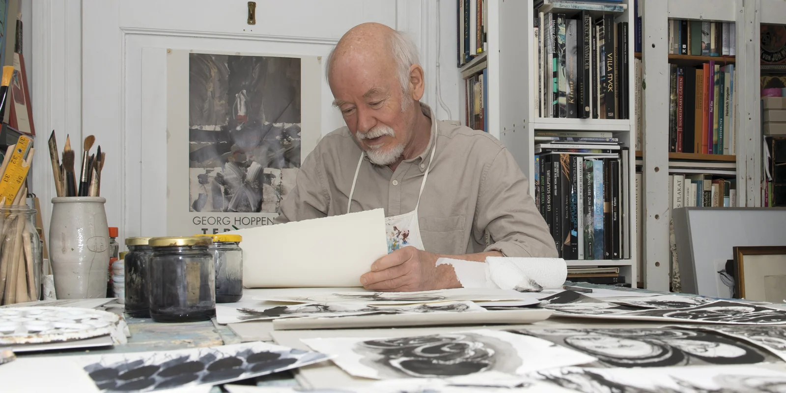 Georg Hoppenstedt sitzt an einem Tisch in seinem Atelier und betrachtet mehrere Bögen mit Zeichnungen.