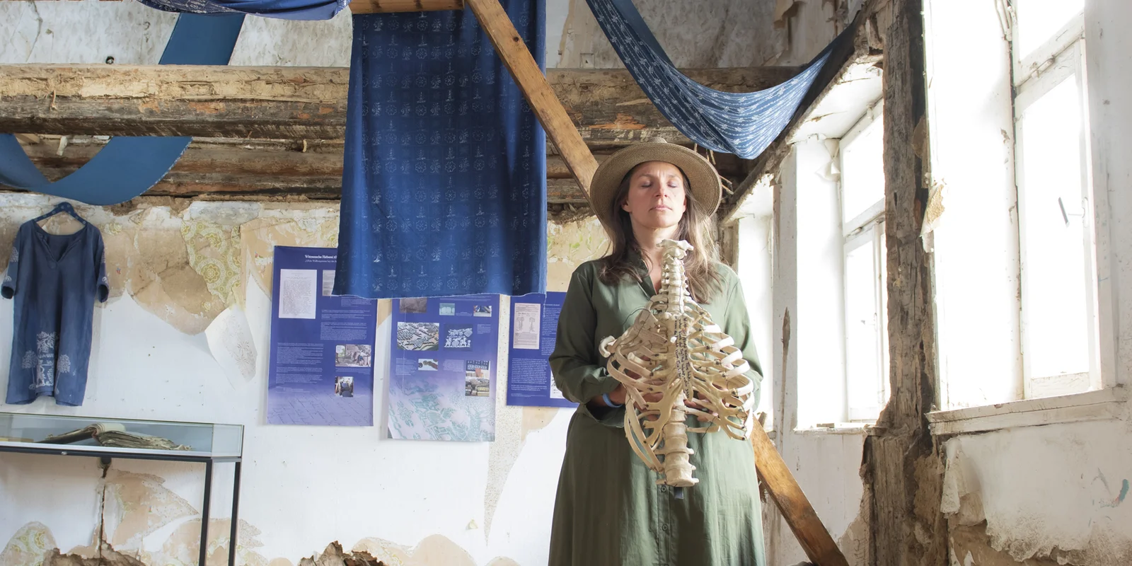 Patricia Magdalene Keil hält den Torso eines Skeletts in den Händen, ihre Augen sind geschlossen. Im Hintergrund ein Fachwerkhaus mit vielen Blaudruck-Stoffen, welche über die Balken gehängt sind.