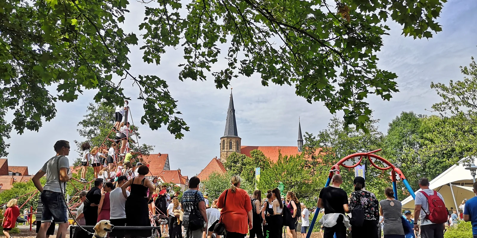 Menschen stehen im LNS-Park in Duderstadt und blicken auf die Freilichtbühne. Im Hintergrund ist ein Klettergerüst zu sehen, im oberen Bildbereich Blätter eines Baumes.